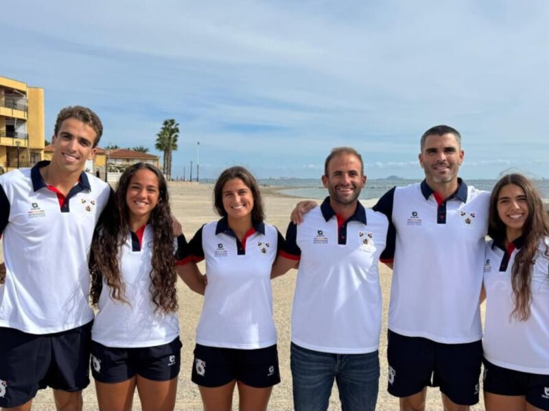 Estudiantes de una escuela deportiva posan en la playa, con uniformes blancos y rojos.