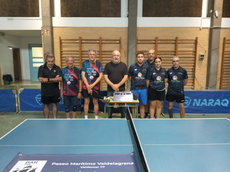 Equipo de tenis de mesa posando frente a una mesa de ping pong en un gimnasio.