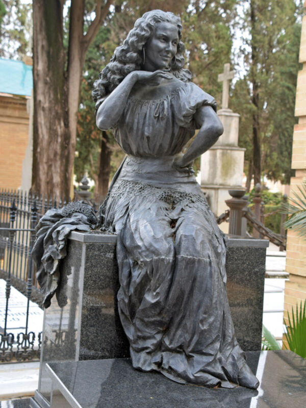 Estatua de bronce de una mujer sentada en un monumento, con cabello largo y vestida con un vestido antiguo. La escultura se encuentra en un cementerio rodeada de árboles y otros monumentos.