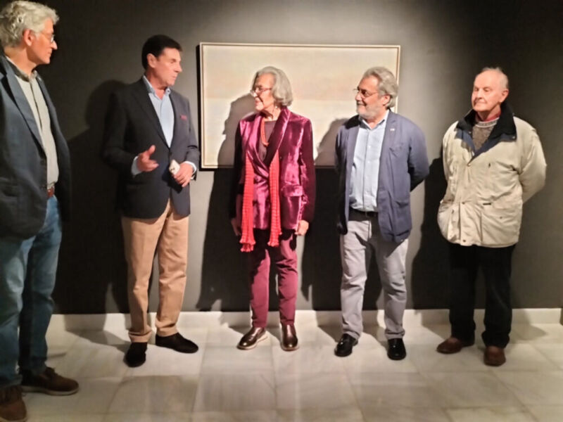 Un grupo de personas posa frente a una pintura en un museo. La mujer central lleva un traje rojo brillante y zapatos negros, mientras que los hombres a su izquierda y derecha están vestidos con chaquetas de lana. La pintura en el fondo muestra un paisaje soleado.