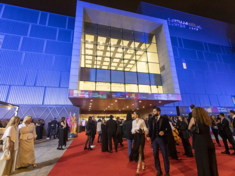Fotografía nocturna de una alfombra roja frente a un edificio moderno con luces proyectadas. Personas vestidas formalmente caminan por el camino de honor hacia la entrada principal del edificio. La iluminación y los colores contrastantes hacen que el evento parezca elegante y sofisticado.