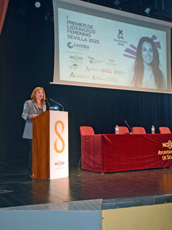 A una mujer en un escenario, posiblemente dando un discurso o presentando algún reconocimiento. En el fondo, se ve una pantalla con el título "Premios de Liderazgo Femenino Sevilla 2025" y varios patrocinadores. La escena se desarrolla en un evento formal, probablemente relacionado con la celebración de premios o reconocimientos.