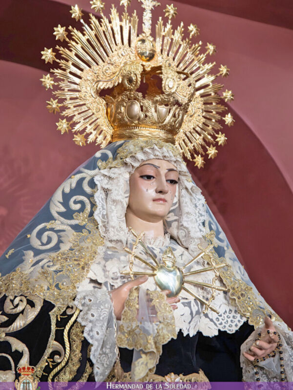 Imagen de una Virgen María vestida con un manto negro y dorado, portando una corona de oro y un halo estrellado. La Virgen sostiene el Sagrado Corazón en su mano izquierda, y su rostro está cubierto por una capa blanca con detalles dorados. La imagen se encuentra en un contexto religioso, posiblemente dentro de una iglesia o templo.