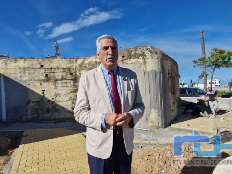 Un hombre en traje blanco y corbata roja posa frente a un edificio militar de estilo clásico, con una estructura circular y murallas. El cielo es azul claro con pocos nubes, y hay una palmera en el fondo. La imagen es parte de un reportaje de la televisión municipal de Chipiona, como se puede ver en el logo.