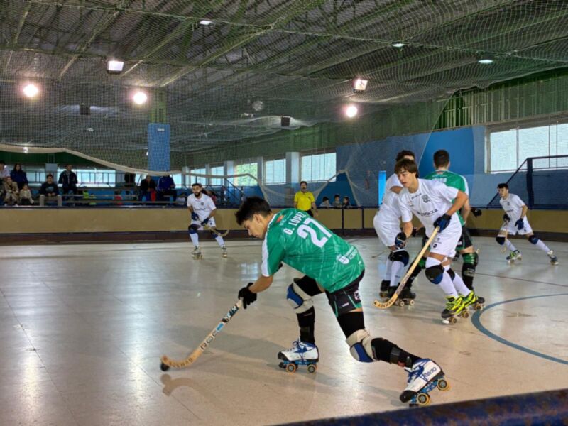 Jugadores en un campo de hockey sobre patines, jugando en una cancha interior con una pista azul y un techo de metal.