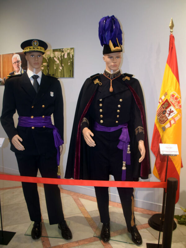 Dos modelos de uniformes militares, uno con un sombrero y otro con una capa, frente a una bandera española.