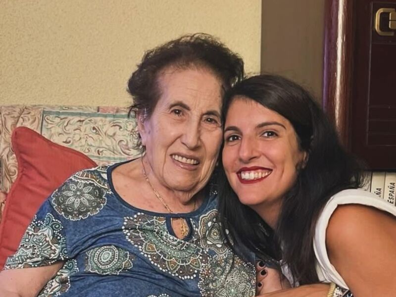 Una abuela sonriente y una niña joven también sonríen, sentadas en un sofá con cojines coloridos. La abuela lleva una blusa azul con detalles en verde y la niña usa un top blanco.
