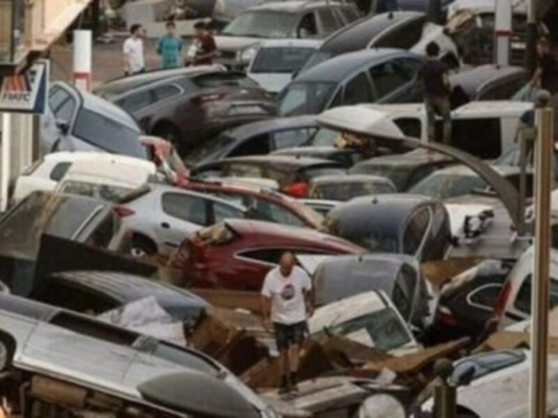 Imagen muestra un aparcamiento lleno de coches estacionados en una calle urbana. La imagen está tomada desde un ángulo que muestra varios vehículos aparcados en diferentes direcciones. La iluminación sugiere que es de día, con sombras indicando la presencia de un sol brillante. En el fondo se pueden ver edificios y una señal de tráfico amarilla.