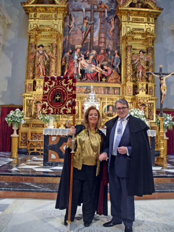 Dos personas posan en un altar de iglesia, rodeados por flores y una imagen religiosa. La escena es formal con vestimentas elegantes, destacando la belleza del entorno histórico.