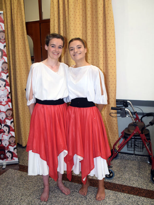 Dos personas vestidas con vestidos blancos y falda roja posan frente a cortinas amarillas. A la derecha, se ve un andador rojo con ruedas y una cesta.