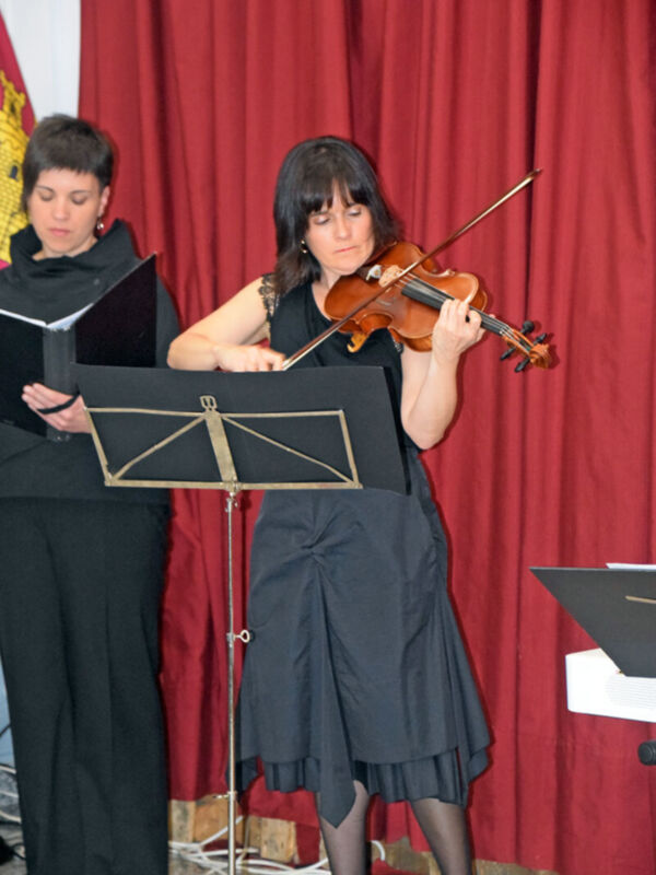 Una mujer toca el violín mientras otra mira un libro de partituras. Fondo rojo con cortinas y piano blanco.