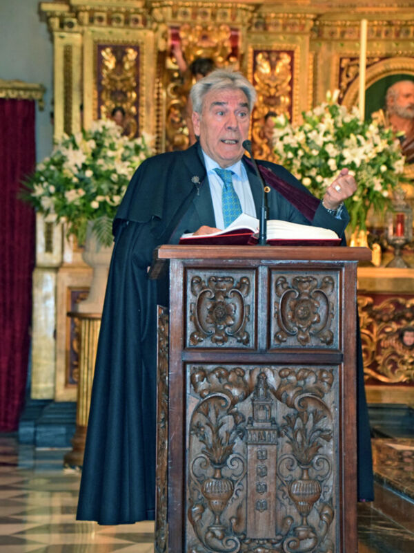 Un hombre en un hábito religioso está hablando frente a una pila alta con flores y un altar en el fondo.